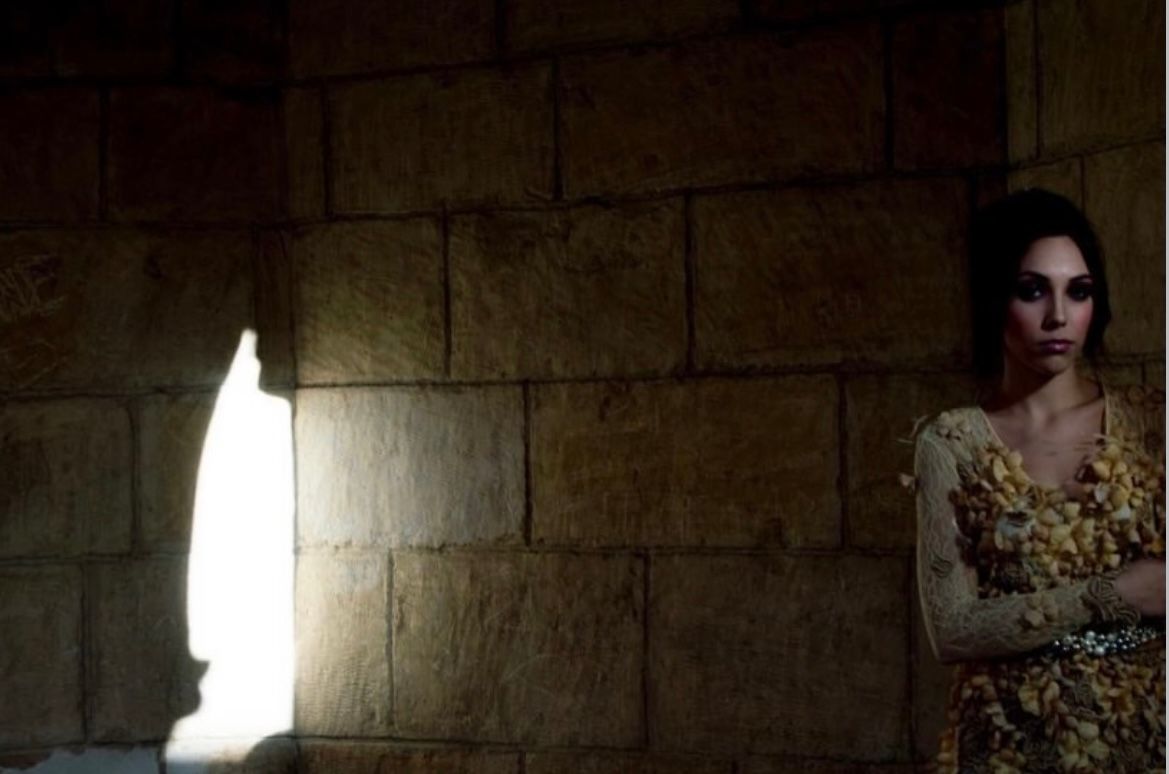 Modelo con vestido de encaje dorado junto a un muro de piedra atravesado por la luz